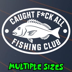 May include: A white oval vinyl decal featuring a central illustration of a fish. The text CAUGHT F*CK ALL is arched above the fish, and the text FISHING CLUB is arched below. Two small white circles are positioned on the left and right sides.