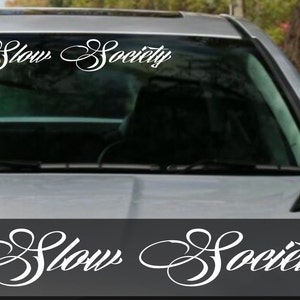 May include: A white vinyl decal featuring the words "Slow Society" in a decorative script. The decal is applied to a car's windscreen. The car is silver, and the background is blurred.
