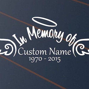 May include: White decal with the text "In Memory of" above "Custom Name" and the years "1970 - 2015". The design includes angel wings and a halo on a dark blue background.