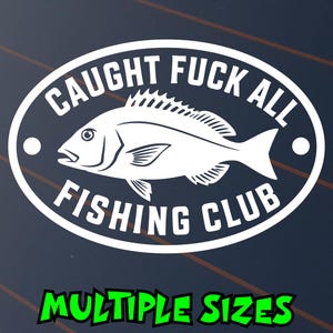 May include: A white oval vinyl decal featuring a central illustration of a fish. The text CAUGHT FUCK ALL is arched above the fish, and the text FISHING CLUB is arched below. Two small white circles are positioned on the left and right sides.