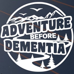 May include: White circular graphic with the text "ADVENTURE BEFORE DEMENTIA". The design includes mountain silhouettes, birds, and stylized trees. The text is in a bold, rounded font.