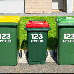 May include: Three green wheelie bins with yellow, red, and green lids. Each bin displays the address "123 APPLE ST" in white text. The bins are positioned on a paved area in front of a lawn.