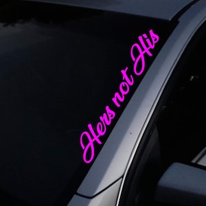 May include: A car window sticker featuring the words "Hers not His" in a vibrant pink, cursive script. The sticker is affixed to the dark glass of a car window, with the vehicle's frame in view.