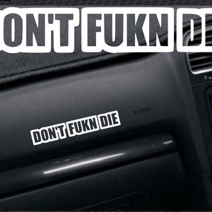 May include: A black car dashboard featuring a white sticker with the text "DON'T FUKN DIE" in bold, capital letters. The sticker is positioned on the dashboard. The image is captured from a slightly oblique angle.