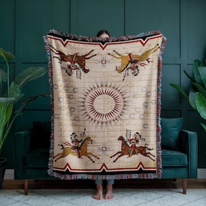 Op de afbeelding: Een geweven deken met een crèmekleurige achtergrond en een Native American ontwerp met vier paarden en ruiters. Het ontwerp is omgeven door een cirkel van rode en witte pijlen en een zwarte en rode rand. De deken heeft een franje rand.