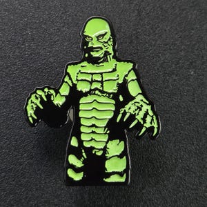 Puede incluir: Pin de esmalte verde y negro con una criatura de la laguna negra. La criatura tiene un cuerpo verde escamoso y una expresión amenazante.
