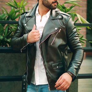 Può includere: Un uomo che indossa una giacca di pelle nera con una camicia bianca sotto. Indossa jeans blu e occhiali da sole neri.