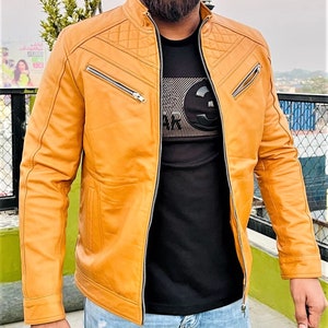 Puede incluir: Una chaqueta de cuero marrón claro con un diseño acolchado en los hombros y cierre de cremallera. La chaqueta tiene varios bolsillos y un cuello de pie.