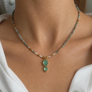 Puede incluir: Un collar con una cadena delicada de pequeñas cuentas de color verde apagado, intercaladas con espaciadores cilíndricos dorados y cuentas blancas. El colgante consta de dos piedras ovaladas de color verde claro translúcido en oro.
