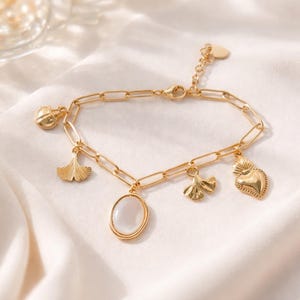Peut inclure: Bracelet doré à breloques avec plusieurs pendentifs. Les breloques comprennent une coccinelle, une feuille de ginkgo, un pendentif ovale avec une pierre blanche, un cœur et une petite feuille. Le bracelet est sur une surface blanche et soyeuse.
