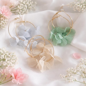 May include: Three pairs of hoop earrings with floral accents in pastel shades: light blue, cream, and mint green. The earrings feature gold-coloured hoops and delicate fabric flower details. They are displayed on a white fabric surface.