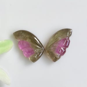 May include: Two carved gemstone pieces shaped like butterfly wings, featuring a translucent olive-green outer edge and a vibrant, deep pink inner section with carved ridges, displayed side-by-side against a plain, neutral white background.