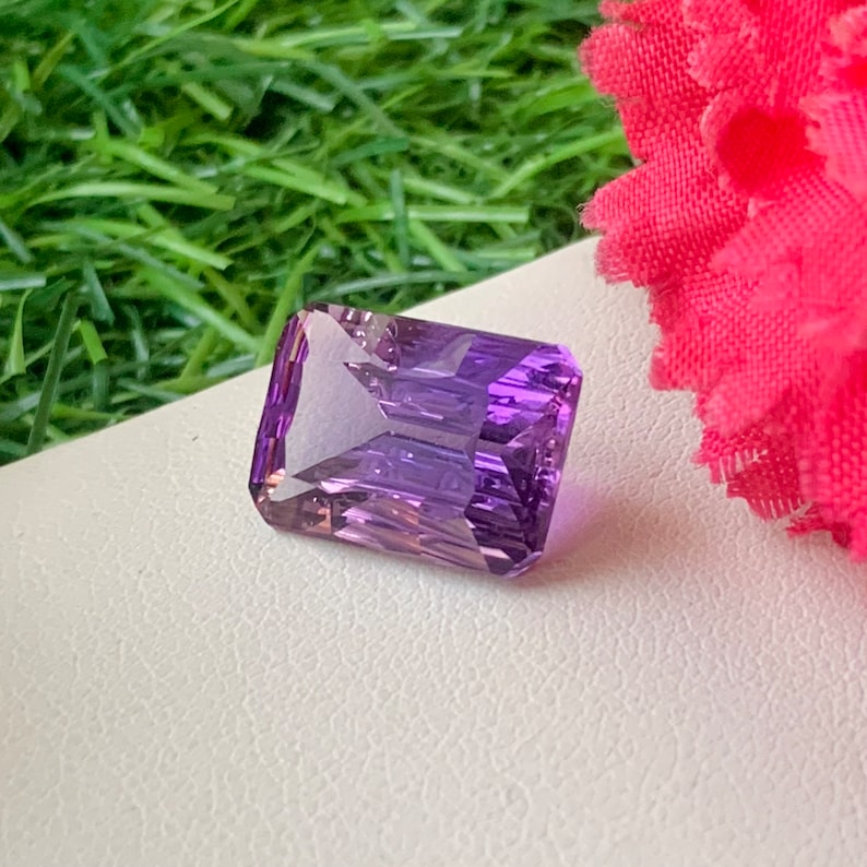 May include: A large, rectangular, purple amethyst gemstone with a faceted cut. The gemstone is on a white surface.