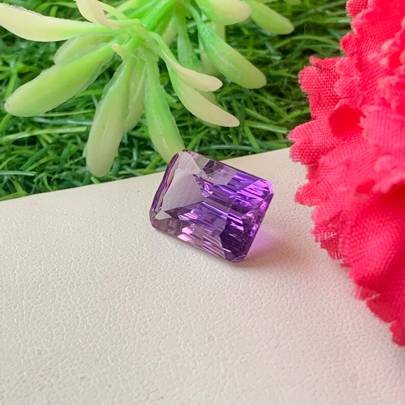 May include: A vibrant purple gemstone with a rectangular emerald cut. The gemstone is set against a white background.