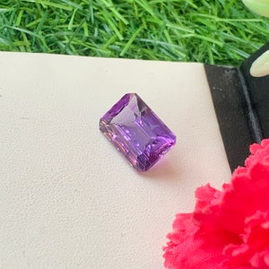 May include: A vibrant purple amethyst gemstone with a rectangular emerald cut. The gemstone is set against a white background.