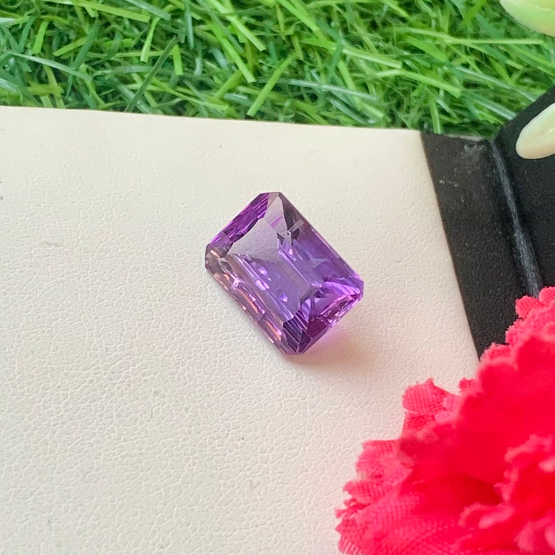 May include: A vibrant purple amethyst gemstone with a rectangular emerald cut. The gemstone is set against a white background.