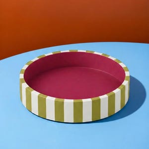 May include: A round tray featuring a deep magenta interior and a vertical striped exterior pattern in alternating olive green and white. The tray has a low, straight wall and a flat base, presenting a clean, geometric design with a smooth finish.