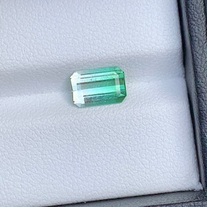 May include: A green and blue gemstone with a rectangular shape. The gemstone is set in a white box.