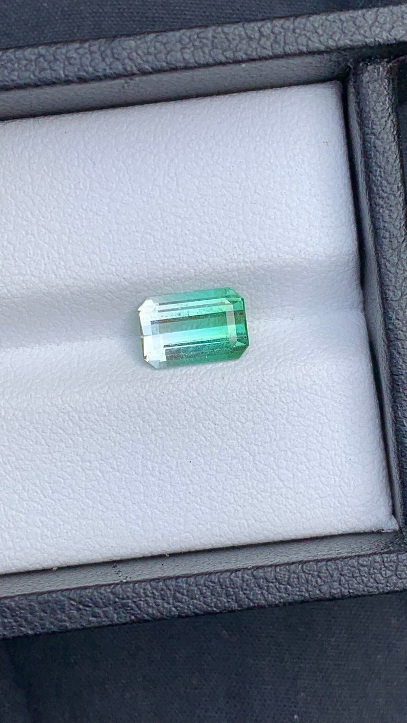 May include: A green and blue gemstone with a rectangular shape. The gemstone is set in a white box.