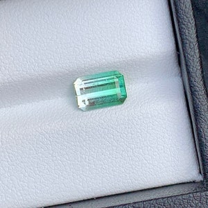 May include: A light green and dark green emerald cut gemstone in a white box.