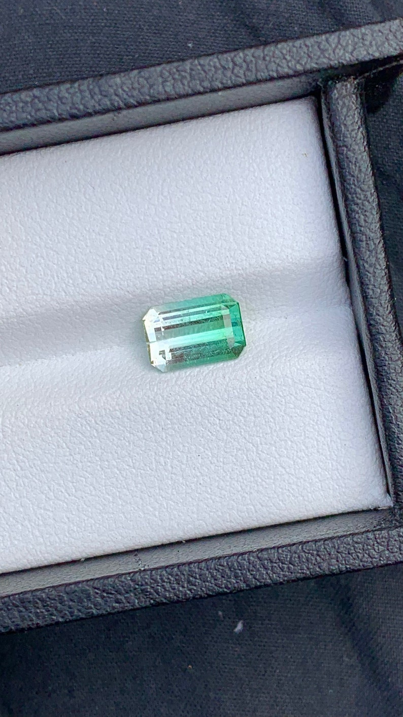 May include: A light green and dark green emerald cut gemstone in a white box.