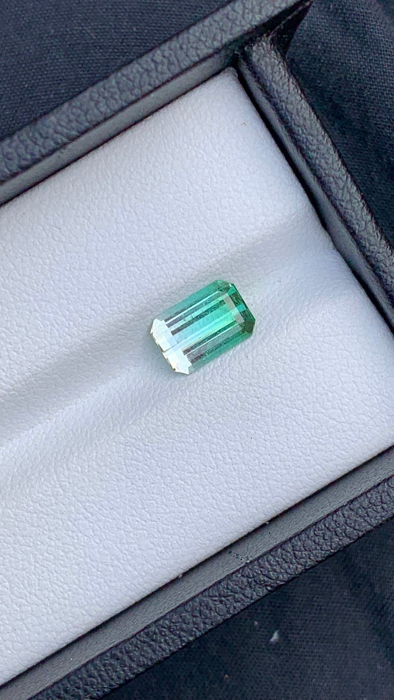 May include: A green and blue gemstone with a rectangular shape, set in a white velvet box.