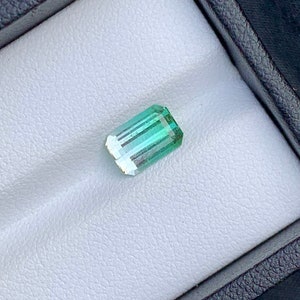 May include: A light green emerald gemstone with a rectangular shape, set in a white velvet box.