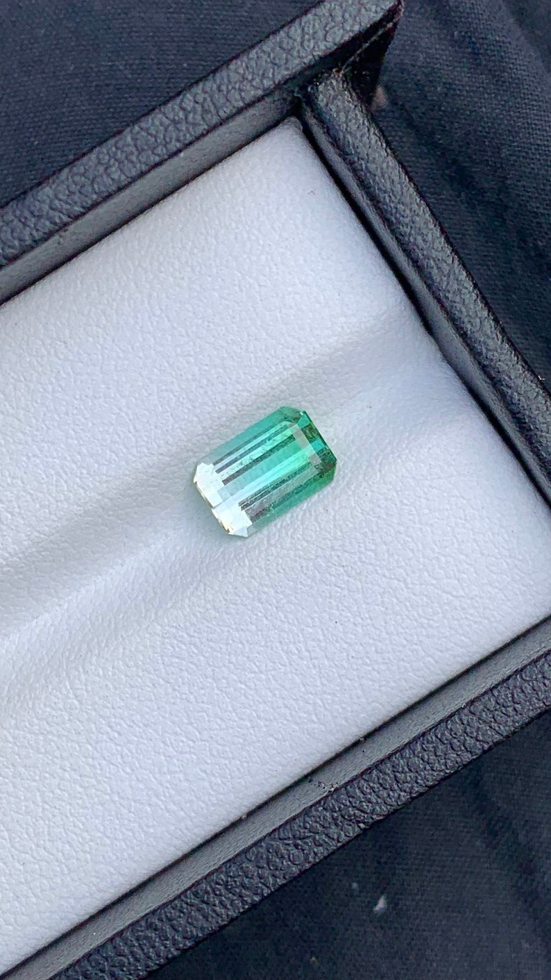 May include: A light green emerald gemstone with a rectangular shape, set in a white velvet box.