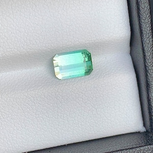 May include: A light green gemstone with a rectangular shape, set in a white display box.