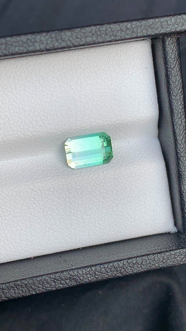 May include: A light green gemstone with a rectangular shape, set in a white display box.