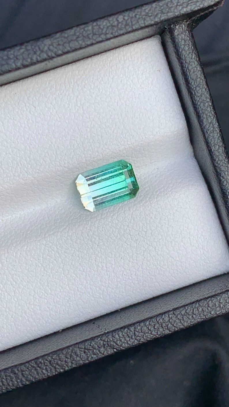 May include: A green and blue gemstone with a rectangular shape, set in a white box.