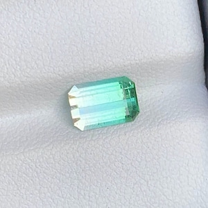 May include: A green and blue gemstone with a rectangular shape. The gemstone has a light green color on one side and a darker green color on the other side.