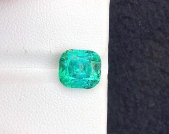 May include: A vibrant, cushion-cut emerald gemstone. The gem is a bright, vivid green color and appears to be faceted. The emerald is set against a white background.