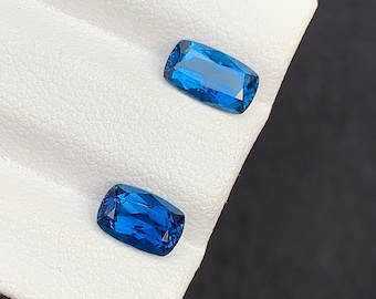 May include: Two blue gemstones, cut in a rectangular shape, are displayed on a white surface. The gemstones are faceted and have a deep blue color.