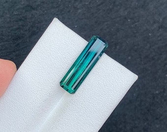 May include: A green and blue gemstone with a rectangular shape and a faceted cut. The gemstone is on a white surface.