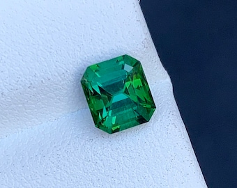 May include: A vibrant green gemstone with a square emerald cut. The gemstone is set on a white background.
