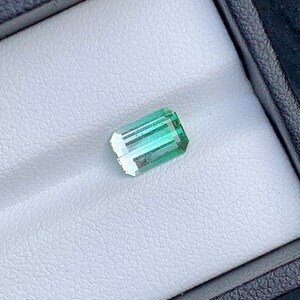 May include: A light green emerald gemstone with a rectangular shape, set in a white velvet box.