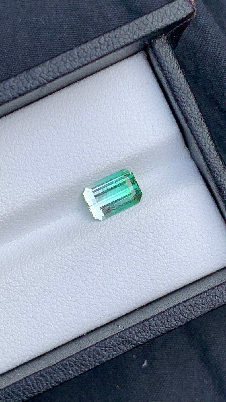 May include: A light green emerald gemstone with a rectangular shape, set in a white velvet box.