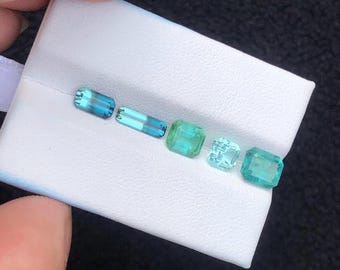 Può includere: Una collezione di cinque gemme in tonalità di blu e verde, esposte su una superficie bianca. Le gemme variano nella forma, inclusi tagli rettangolari e quadrati. Il colore varia dall'acqua marina chiara al verde-azzurro intenso.