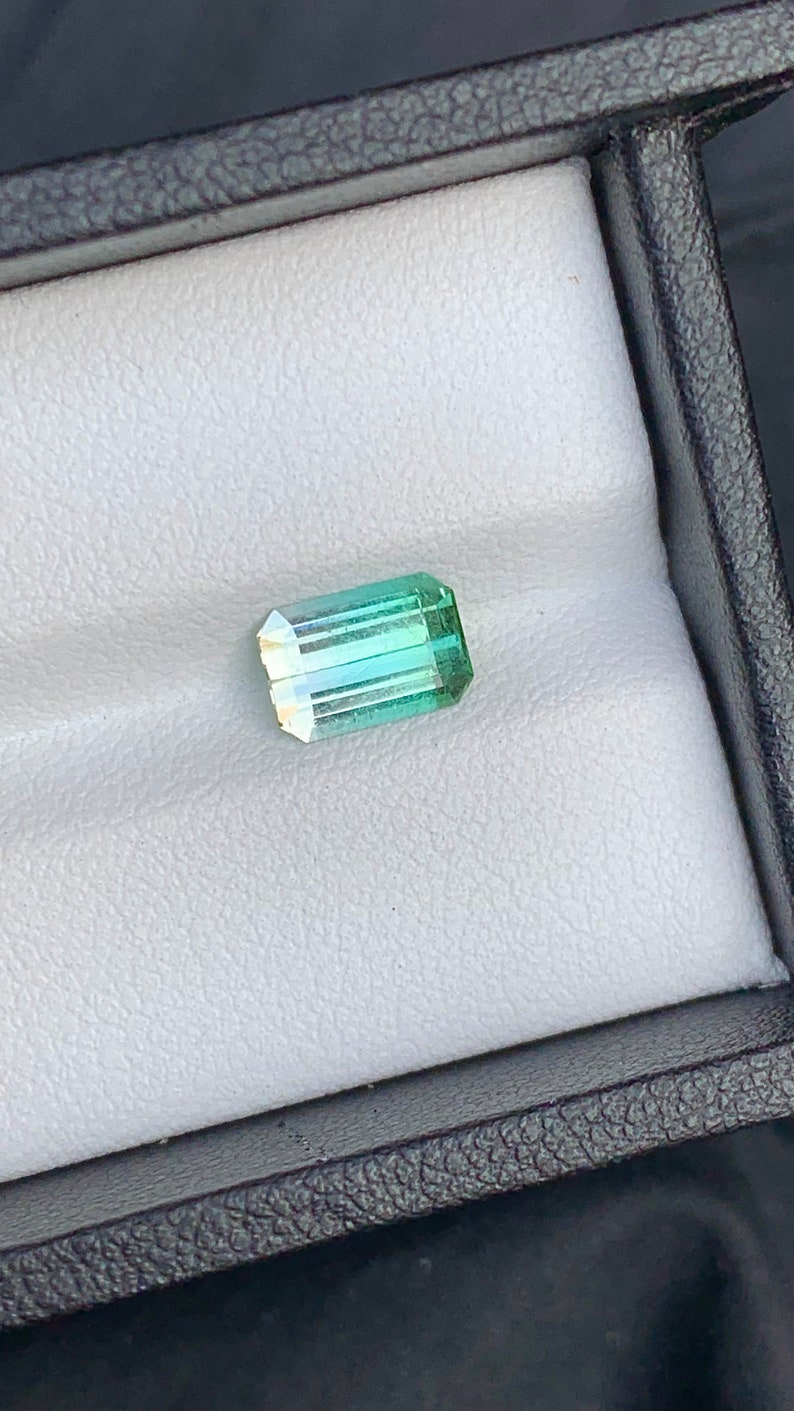 May include: A green and blue gemstone with a rectangular shape, set in a white velvet box.