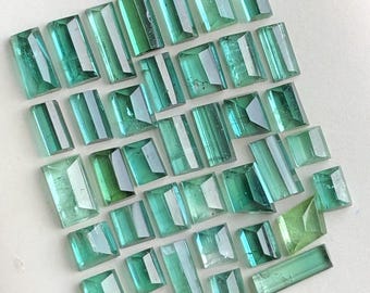 Könnte beinhalten: Eine Ansammlung rechteckiger und quadratischer Edelsteine in verschiedenen Türkis- und Grüntönen. Die Edelsteine sind auf einer weißen Oberfläche angeordnet, um ihre Facetten und Klarheit zu zeigen. Die Edelsteine sind wahrscheinlich für die Schmuckherstellung bestimmt.