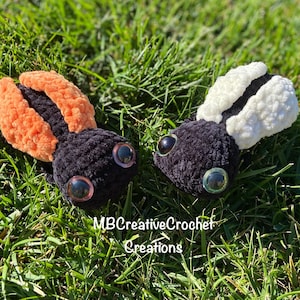 May include: Two handmade crochet bugs with large, googly eyes rest on green grass. One bug has orange wings, the other has white wings. The bugs are black with the text "MBCreativeCrochet Creations" in white.