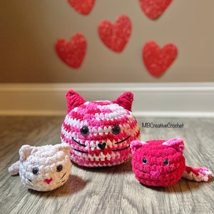 Könnte beinhalten: Drei gehäkelte Katzen-Plüschtiere in Rosa und Weiß, mit schwarzen Augen und einer schwarzen Nase. Die größte Katze ist rosa und weiß mit einem gestreiften Muster. Die beiden kleineren Katzen sind einfarbig rosa und weiß. Die Katzen sitzen auf einem Holzboden mit roten Herzen an der Wand hinter ihnen. Der Text "MBCreativeCrochet" ist im Hintergrund sichtbar.