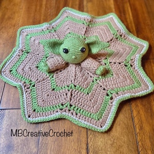 May include: A crocheted baby blanket in the shape of a star with a green crocheted baby toy in the center. The blanket is brown and green with a white border. The toy has large eyes and a small mouth.