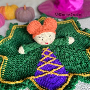May include: A crocheted witch doll with orange hair and a purple dress, lying on a green crocheted blanket with yellow trim. The blanket has a scalloped edge. The doll is holding a green crocheted wand.