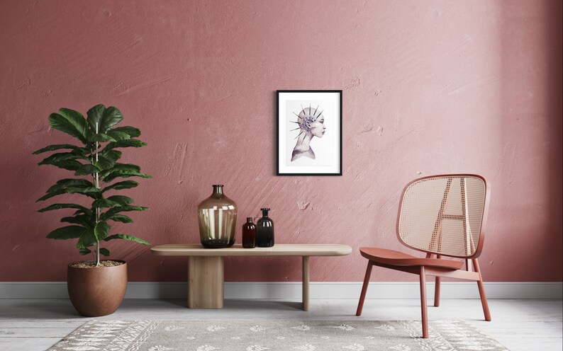 May include: A framed print of a watercolor illustration of a woman's profile with a crown of thorns on her head. The print is hanging on a pink wall in a room with a chair and a plant.