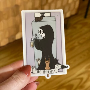 May include: A sticker featuring a tarot card illustration of The Hermit. The card is purple with a white border. The Hermit is depicted as a skeleton wearing a black robe and holding a lantern. Three cats are peeking out from behind the Hermit. The text "IX THE HERMIT" is printed on a white banner at the bottom of the card.