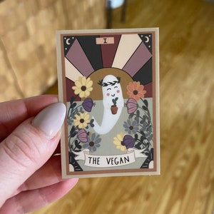 May include: A tarot card with a light green background featuring a white ghost holding a small plant. The ghost has a smiling face and a wreath of flowers on its head. The card is titled "The Vegan" and has a Roman numeral I in the top right corner.