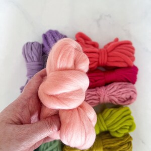 May include: A hand holding a bundle of pink wool yarn. The yarn is surrounded by skeins of yarn in various colors, including purple, red, pink, green, and yellow.