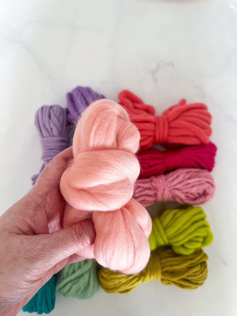 May include: A hand holding a bundle of pink wool yarn. The yarn is surrounded by skeins of yarn in various colors, including purple, red, pink, green, and yellow.